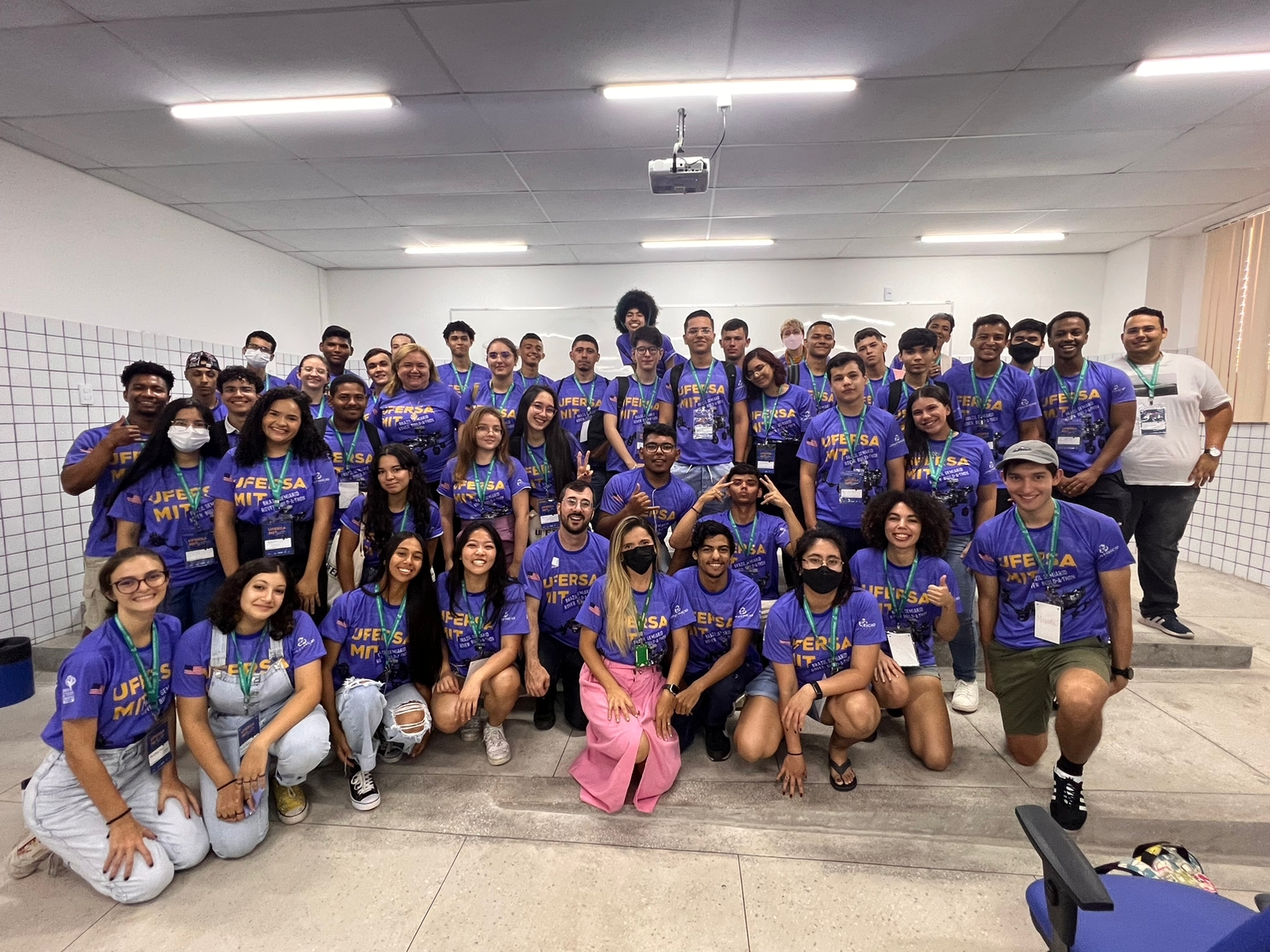 Manuel Morales in a cap and on one knee with 39 high school students in a brightly lit room with tiled walls and cream curtains