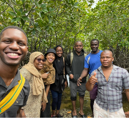 Seven individuals stand and pose for a group photo in a lush green wooded area. 