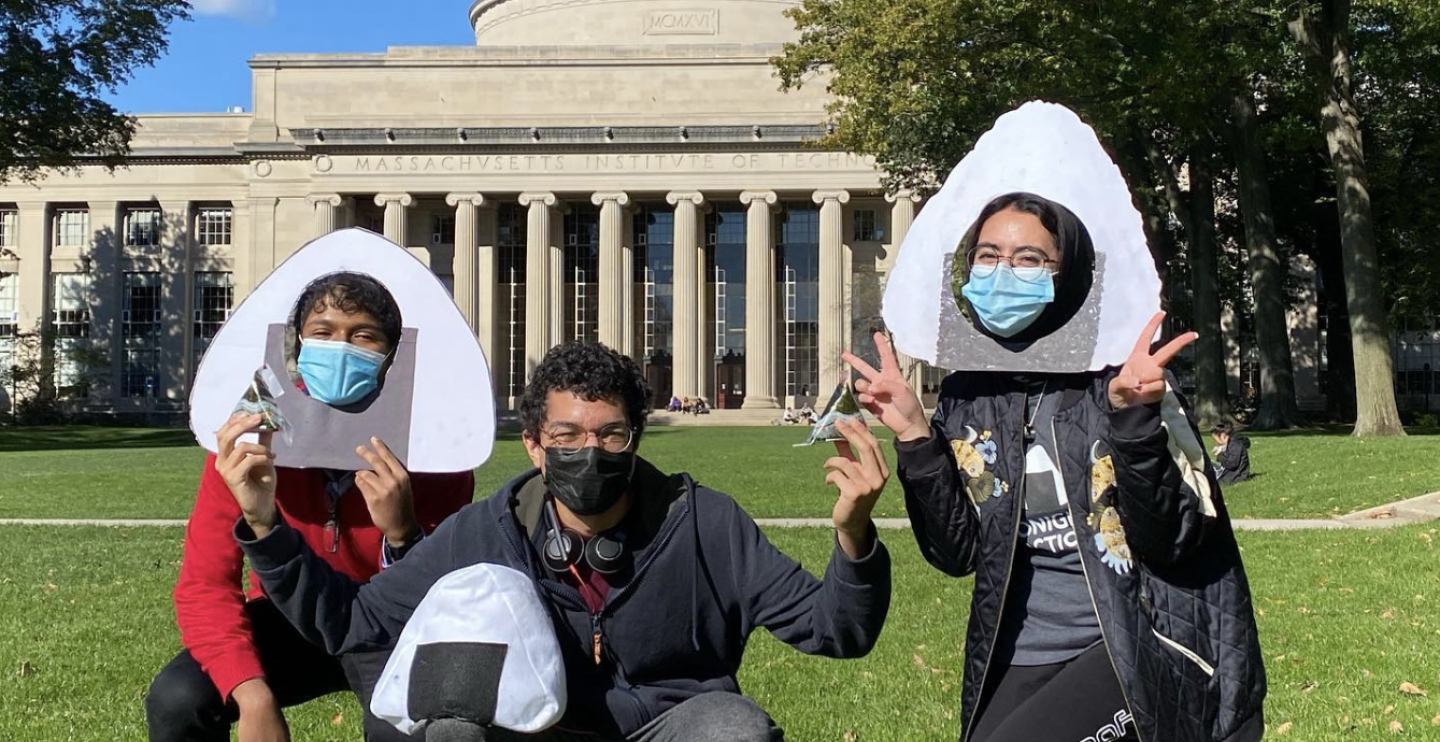 Marwa AlAlawi wearing a onigiri photobooth prop holding two peace signs with both hands crouching down halfway to the right with two other MIT students who's also crouching down on the grass, one wearing a onigiri photobooth prop and another with glasses holding an onigiri rice ball in one hand and rested on his thigh, an onigiri pillow in front of Killian Court at MIT