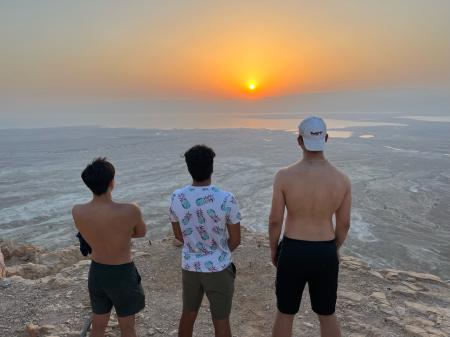 Yajvan Ravan standing in the middle of two other MIT students on top of Masada fortress in Israel, overlooking the Dead Sea and looking out at the sunrise in the horizon