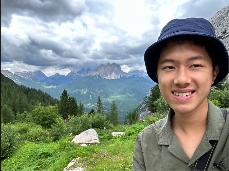 Selfie of David Chang in a blue bucket hat on a cloudy but sunny day with a luscious green forest and mountainous range in the background